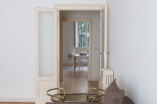 A room with a glass table and chairs, a radiator, and a door leading to a dining area with a table and chairs.