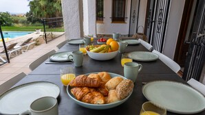 A dining table set with plates, bowls of fruit, croissants, and glasses of orange juice, overlooking a pool and greenery. - Villa 'Ensueño' with Sea View, Wi-Fi and Air Conditioning (Calonge)