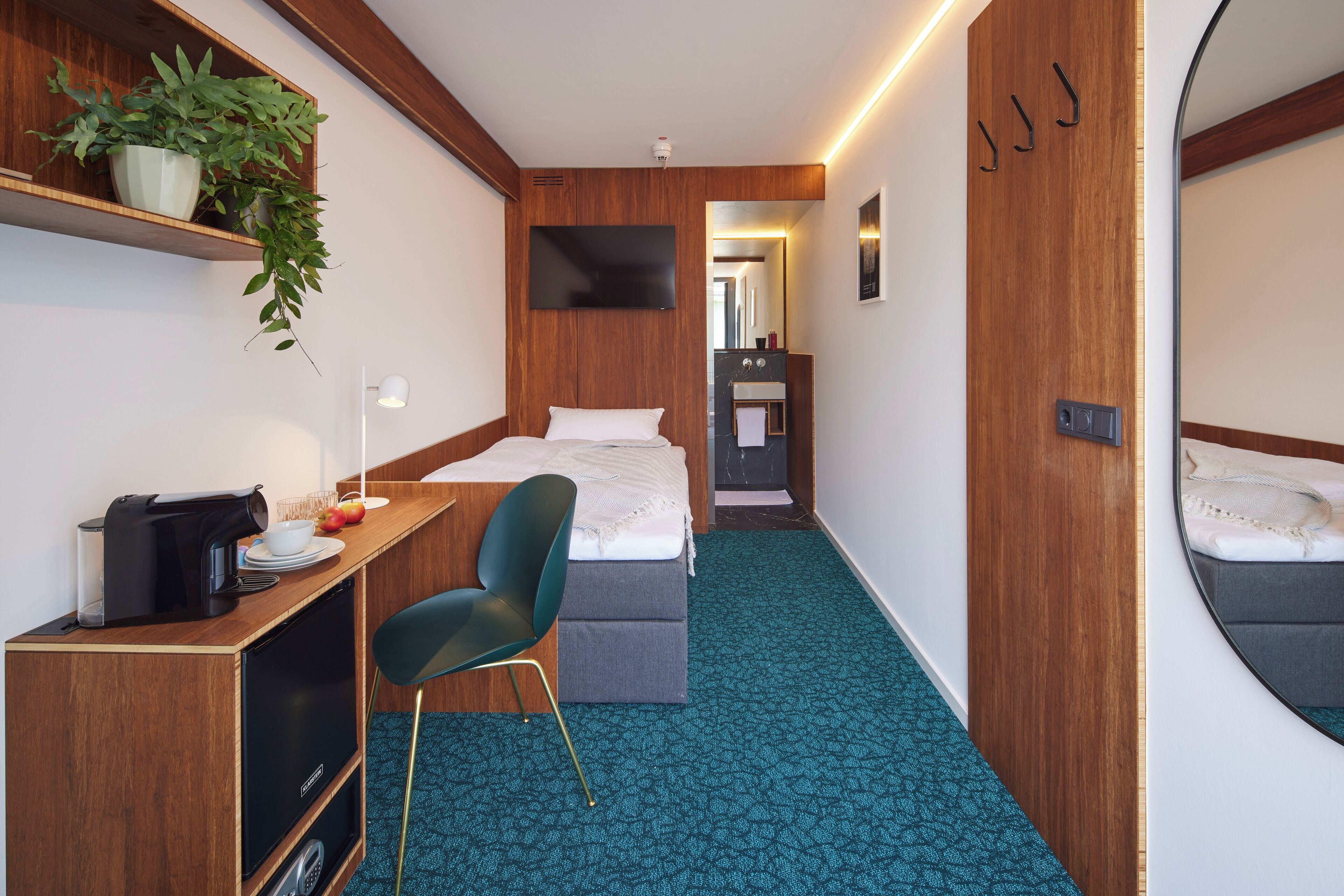 Une chambre d’hôtel compacte comprenant un lit, un bureau avec une chaise, une petite kitchenette, un miroir et une plante en pot.
