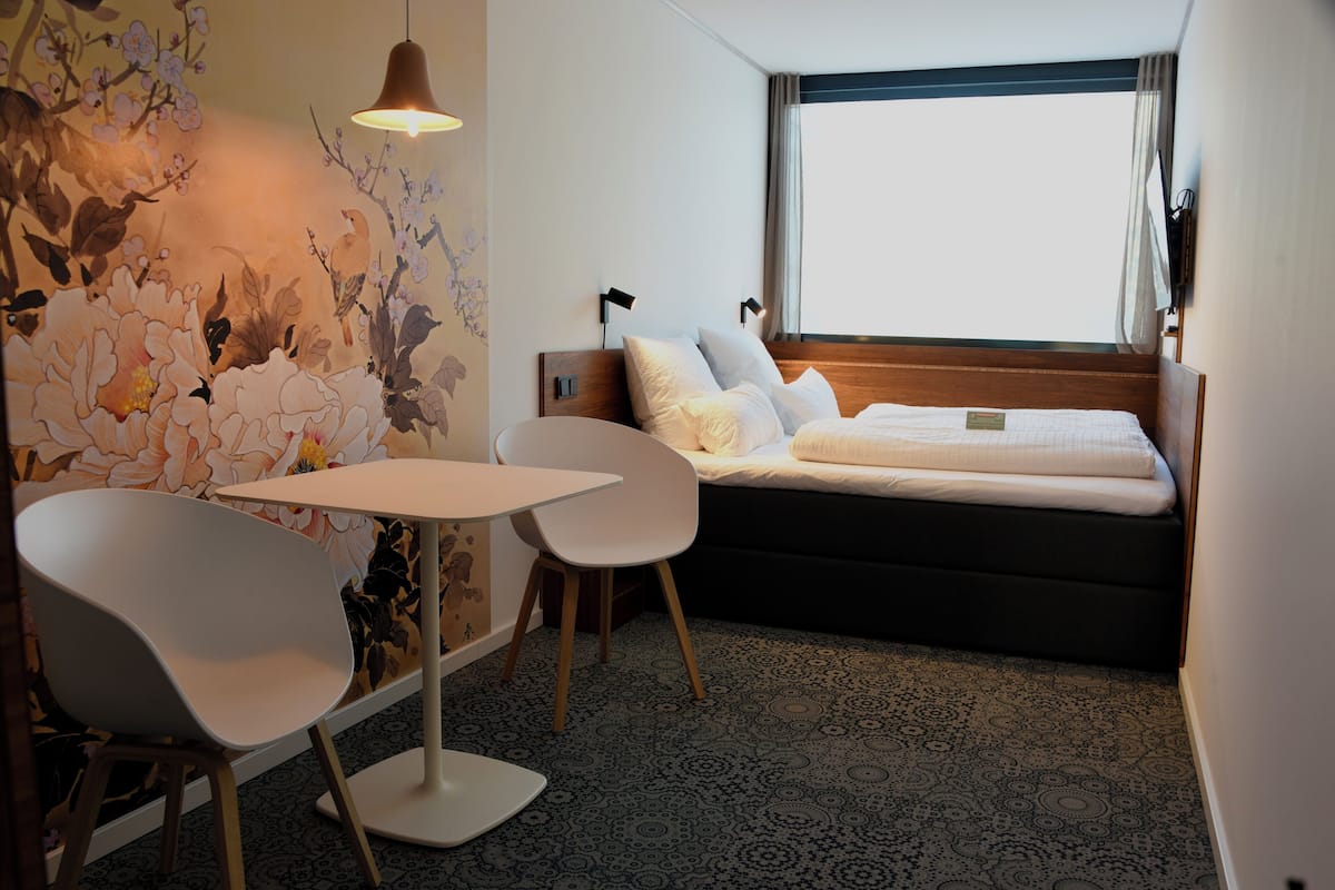 A hotel room with a bed, a table, two chairs, a wall mural, and a window with curtains.