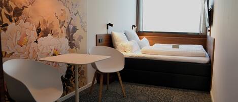 Une chambre d’hôtel avec un lit, une table, deux chaises, une fresque murale et une fenêtre avec des rideaux.