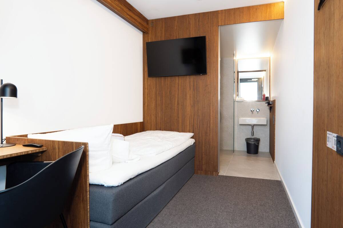 A hotel room with a bed, desk, chair, TV, and a small bathroom with a sink and mirror.