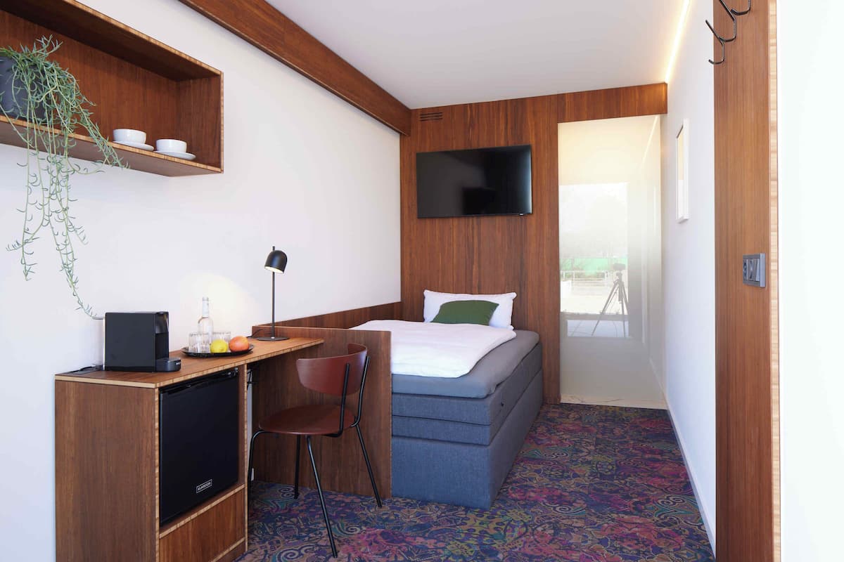 A compact hotel room with a bed, a desk with a computer, a chair, a small fridge, a TV mounted on the wall, and a window with a view.