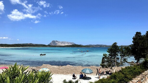 En strand med kristallklart turkost vatten, en stenig kustlinje och ett berg i bakgrunden.
