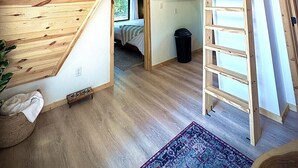 Une chambre de style « loft » avec un plafond en pente en bois, un téléviseur fixé au mur, une échelle menant à une zone de couchage en mezzanine et une petite fenêtre.