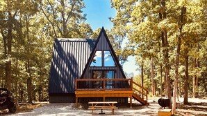 Une cabane avec un toit triangulaire, une terrasse en bois et une table de pique-nique, située dans une zone boisée.