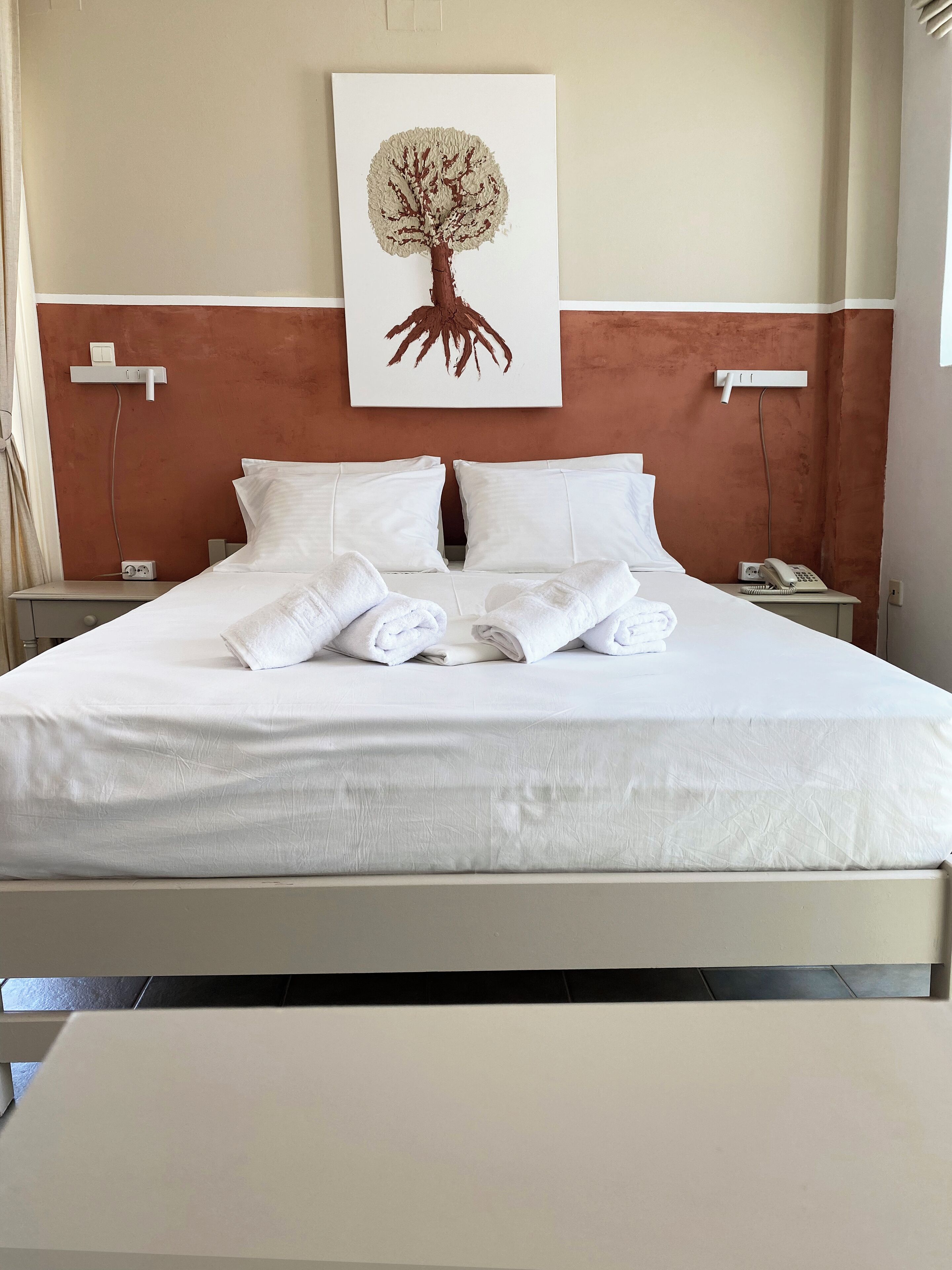 Un lit bien fait, avec du linge de lit et des serviettes blanches, une œuvre d’art représentant un arbre accrochée au mur, et un téléphone sur une table de chevet.