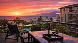 Un balcon avec une table, des chaises, une bouteille de vin et des verres, donnant sur une zone de villégiature bordée de palmiers et offrant une vue sur le coucher du soleil.