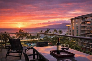 A balcony with a table, chairs, a bottle, and glasses, overlooking a resort area with palm trees and a sunset.