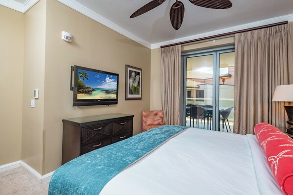 A hotel room with a bed, a TV, a desk, a chair, a balcony with curtains, and a ceiling fan.