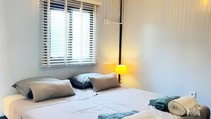Un lit bien fait, avec des oreillers et des serviettes enroulées, une fenêtre avec des stores, une lampe et un luminaire au plafond.
