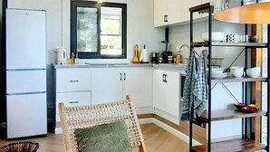 Une cuisine compacte avec des meubles blancs, une chaise en osier dotée d’un coussin, un lampadaire en fibres tressées et une étagère avec de la vaisselle.