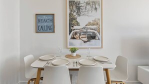 Un comedor con una mesa blanca dispuesta para cuatro, una obra de arte enmarcada encima y una planta pequeña sobre la mesa.