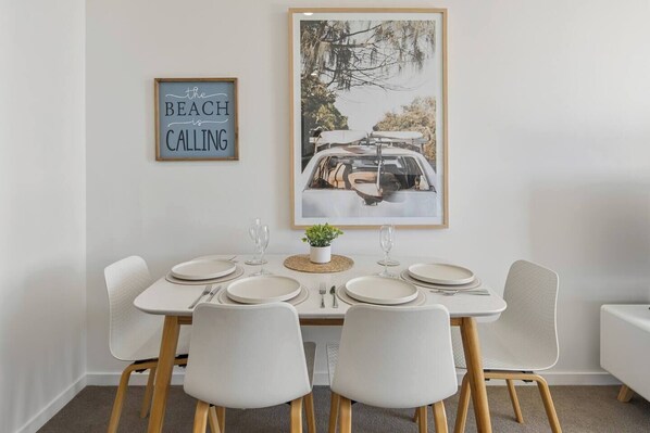 Un comedor con una mesa blanca dispuesta para cuatro personas, un cuadro enmarcado encima y una planta pequeña sobre la mesa.
