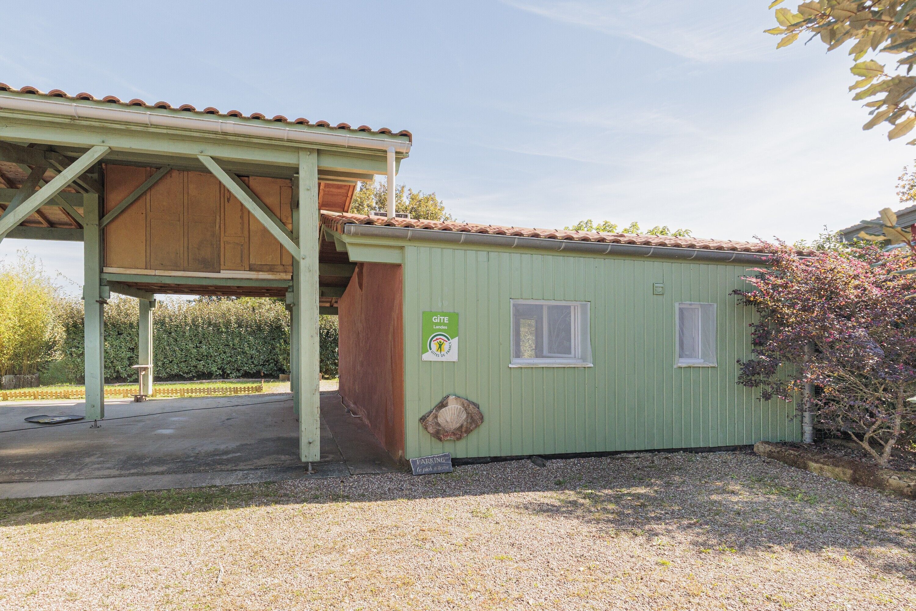 Een klein, laag gebouw met een groene gevel en pannendak. Het heeft een overdekt buitengebied met een houten bankje en een bordje dat een parkeerplaats aangeeft.