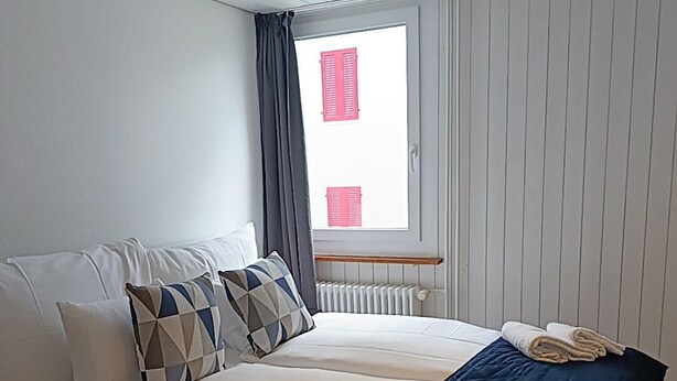 A double bed with white and blue bedding, a geometric patterned pillow, and a dark blue throw blanket. There are towels on the bed and a window with red shutters.