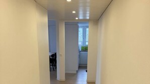 A hallway with wooden flooring and white walls, leading to a kitchen area with a window.