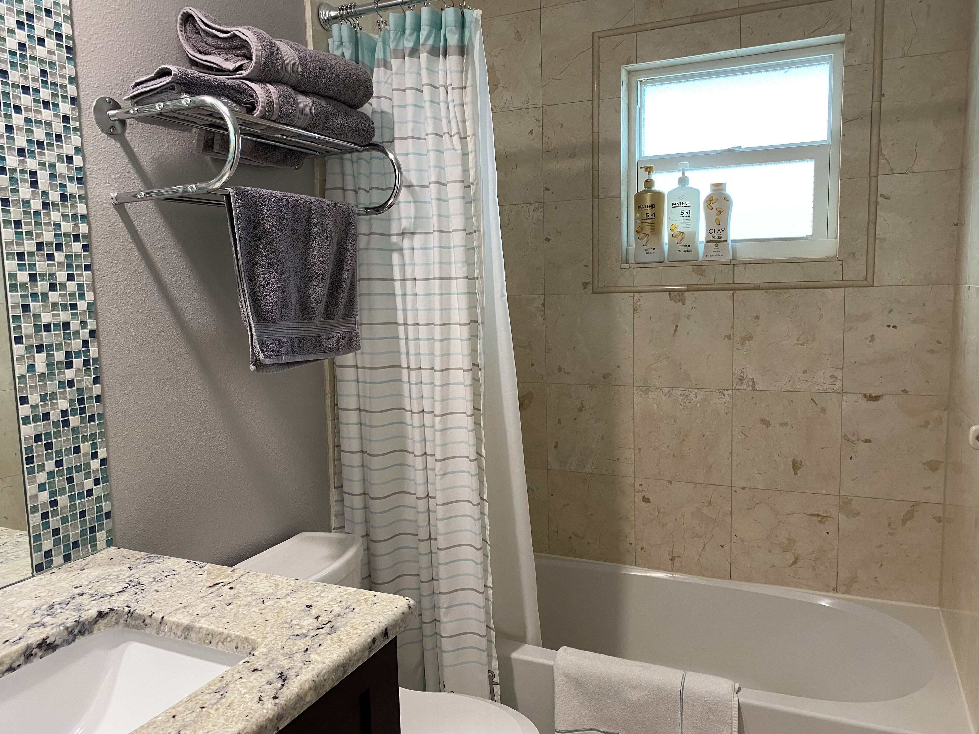 A bathroom with a bathtub, a shower curtain, a towel rack with towels, a vanity with a sink, and a window with toiletries.