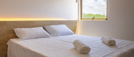 Una cama bien hecha con sábanas blancas y dos toallas enrolladas, un cabecero de madera y una ventana con vistas a la vegetación.