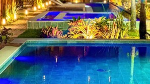 A pool area with a clear view of the pool, surrounding plants, and a building with multiple balconies. - TMH - Essence Condominium in Porto de Pedras/al (Porto de Pedras)