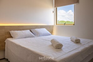 Un lit bien fait, avec du linge de lit blanc et deux serviettes enroulées, une tête de lit en bois et une fenêtre donnant sur un paysage verdoyant.