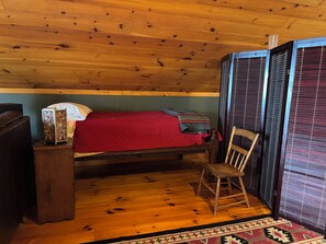 Una habitación de cabaña acogedora con techo de madera, una cama individual con una colcha roja, una silla de madera y una mesita con un adorno.
