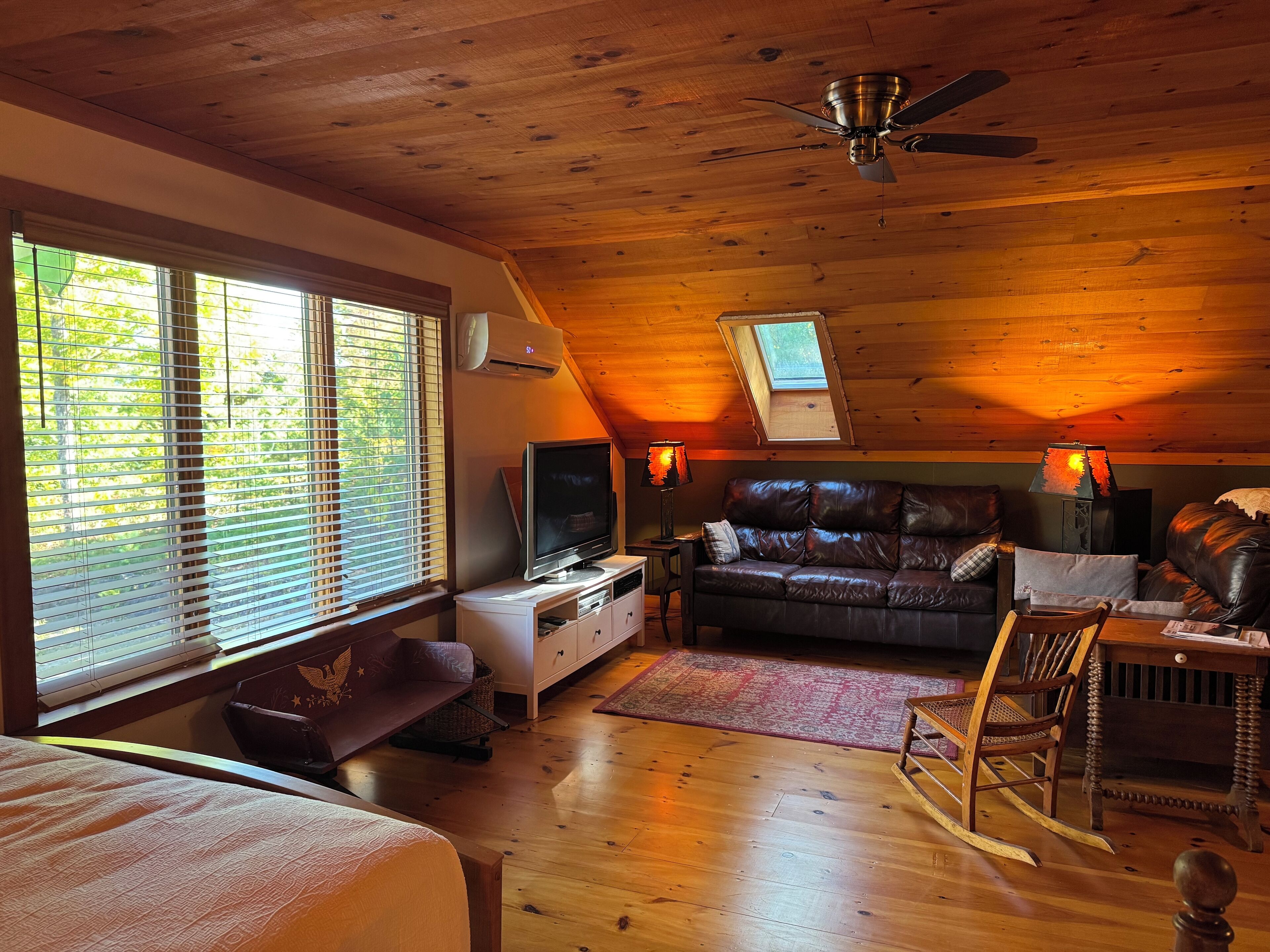 A cozy attic room with a wooden ceiling, a ceiling fan, a TV, a leather sofa, and a skylight.