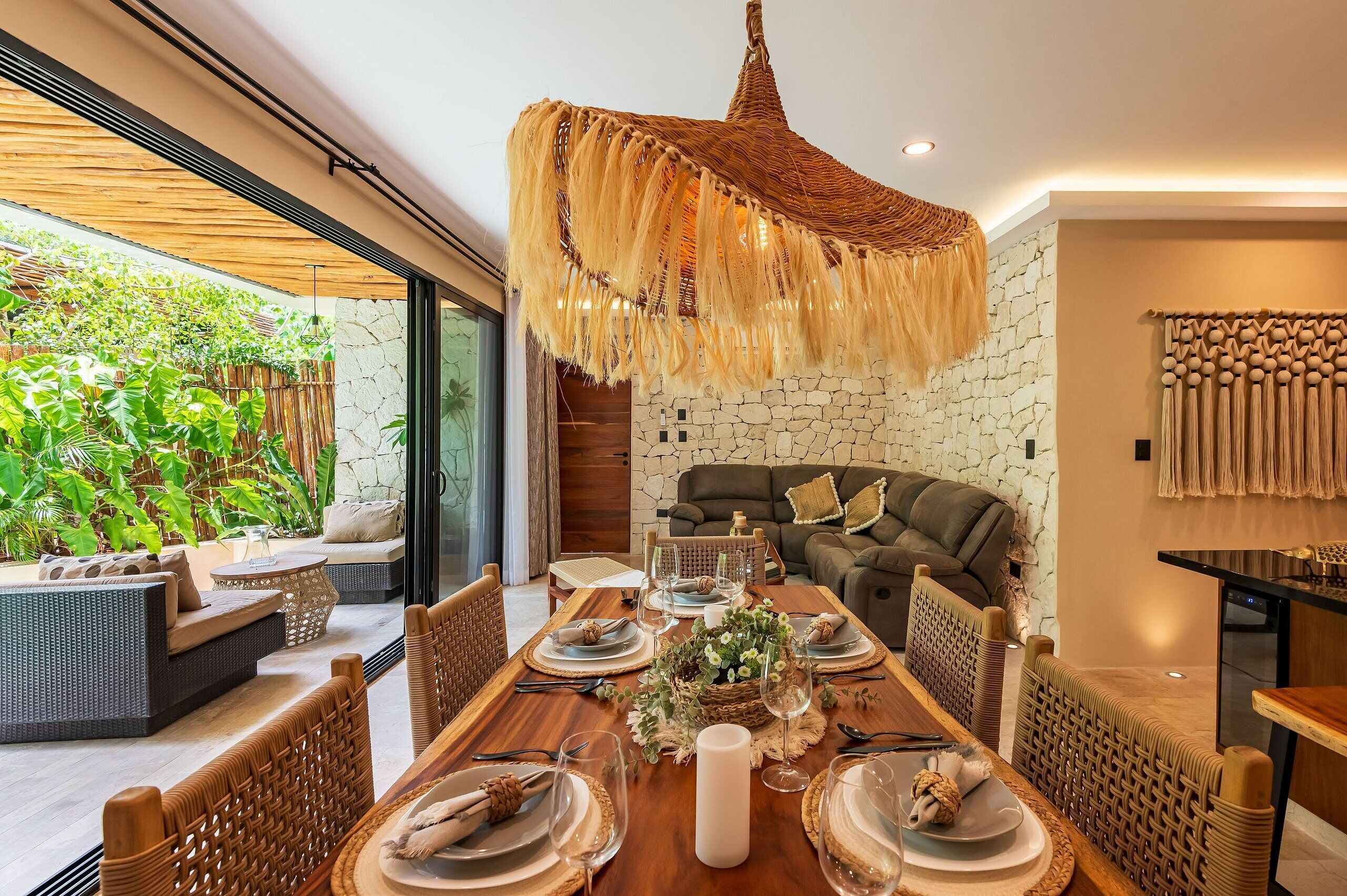 A dining area with a large wooden table set for a meal, surrounded by wicker chairs, a woven hanging light fixture, and a stone wall with a green garden view.
