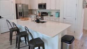 Een moderne keuken met witte kasten, een groot eiland met barstoelen, roestvrijstalen apparatuur en een turquoise koffiezetapparaat.