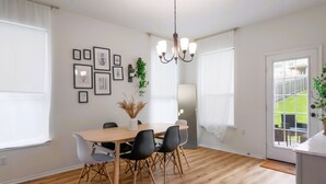Uma sala de jantar com uma mesa de madeira, cadeiras pretas, um lustre, quadros enquadrados e vista através de uma porta de vidro.