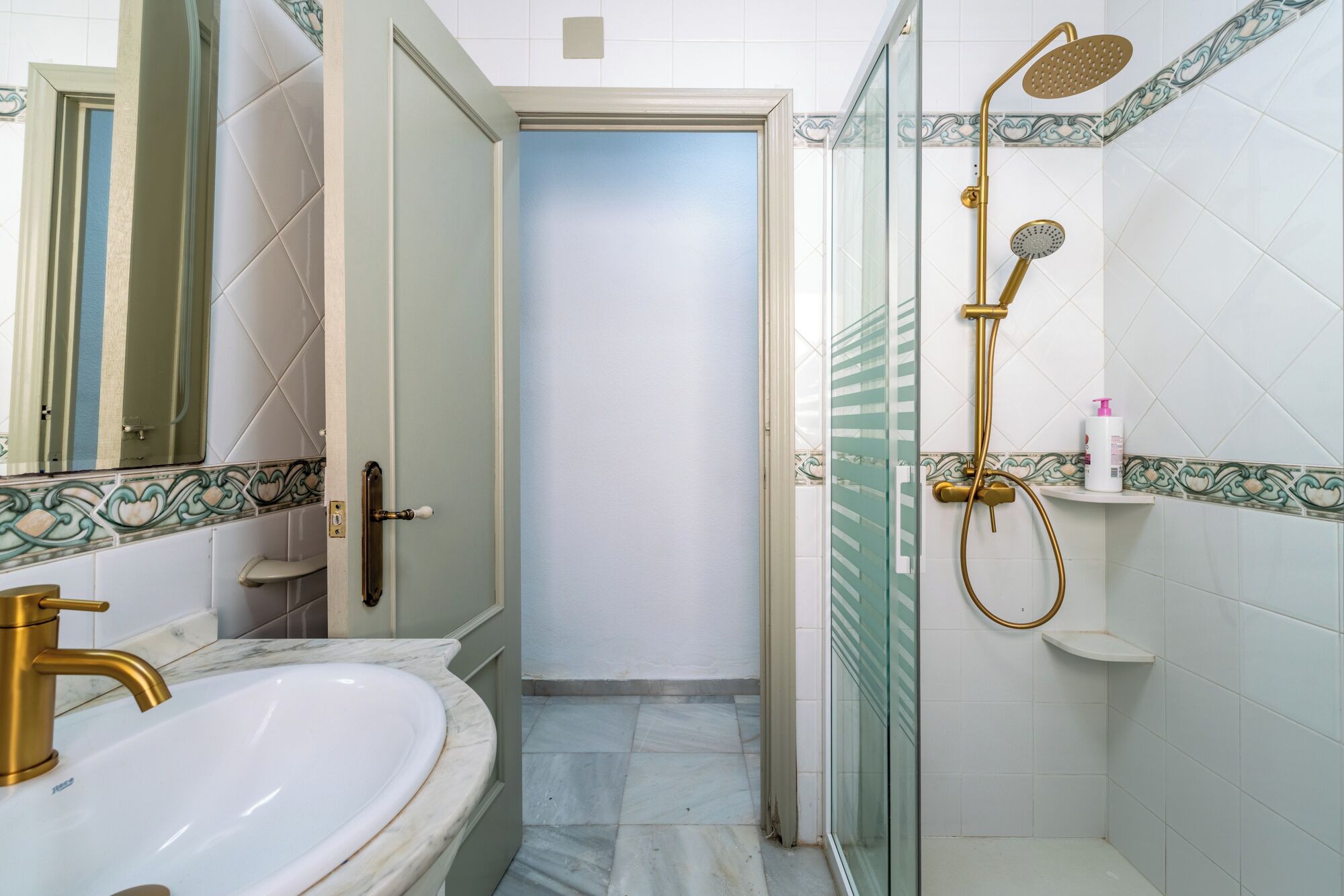 Een badkamer met een wit wastafel, gouden details, een douche met gouden douchetuit en decoratieve tegels.