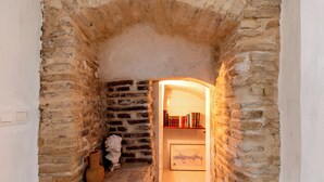 A narrow stone archway leading to a room with a tiled floor, a small shelf with books, and a vase.