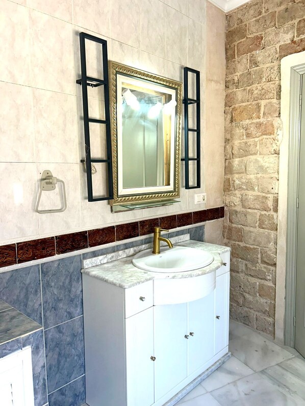 Un baño con lavamanos de mármol, armarios blancos, grifo dorado y espejo con marco dorado.