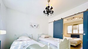 A bedroom with two single beds, a chandelier, a blue door, and a window with blinds.