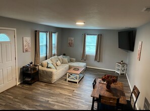 Un salon chaleureux avec un canapé d’angle, une table basse en bois et une table à manger avec des chaises. Un téléviseur à écran plat est fixé au mur, il y a une petite table d’appoint et une fenêtre avec des rideaux.