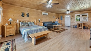 L’intérieur d’une cabane en rondins, avec un plafond en bois, un lit recouvert d’une couverture à motifs, un canapé bleu, un banc en bois et un coin repas avec une table et des chaises.