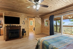 Un dormitorio de cabaña de madera con chimenea, televisor y una ventana grande con vista a un balcón.