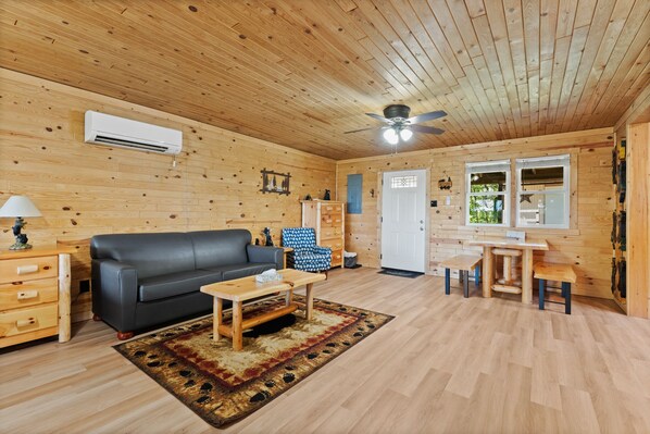 Interior de una cabaña de madera con sofá negro, mesa de centro de madera y comedor con mesa y bancos.