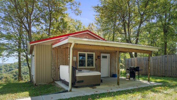 Une petite cabane en bois avec un toit rouge, un jacuzzi et un espace barbecue, le tout entouré d’une clôture en bois et d’arbres.