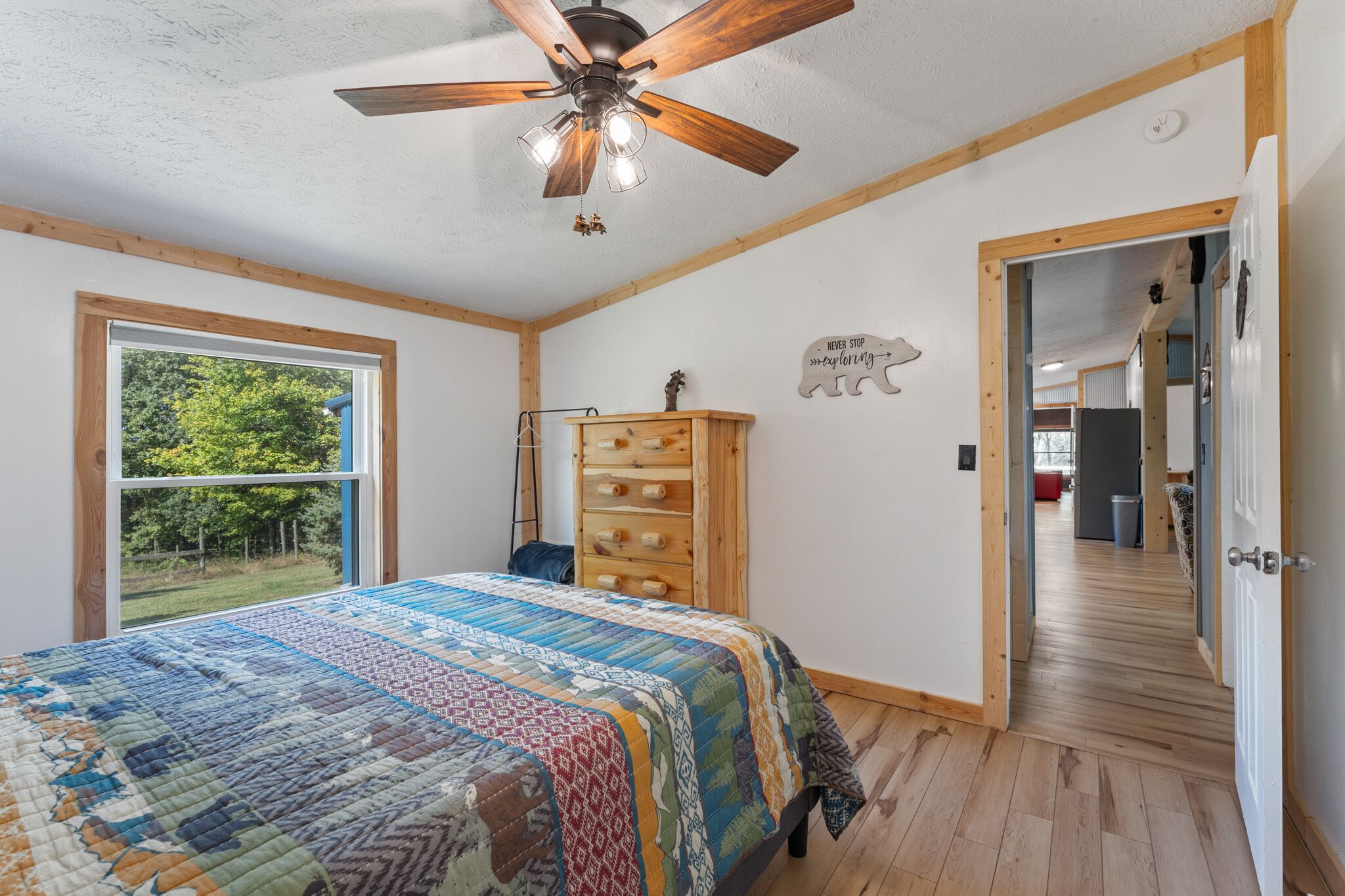 A bedroom with a large bed, a ceiling fan, a window with a view of greenery, a wooden dresser, and a hallway leading to another room.