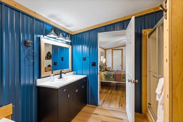 Un baño con un espejo grande, lavamanos y muebles oscuros. Una puerta da a un dormitorio con cama y ventana.