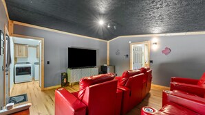 Una sala de estar con sofás de cuero rojo, un televisor de pantalla plana, techo de madera y una cocina visible a través de un umbral abierto.