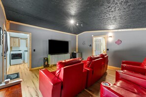 A living room with red leather sofas, a flat-screen TV, a wooden ceiling, and a kitchen area visible through an open doorway.