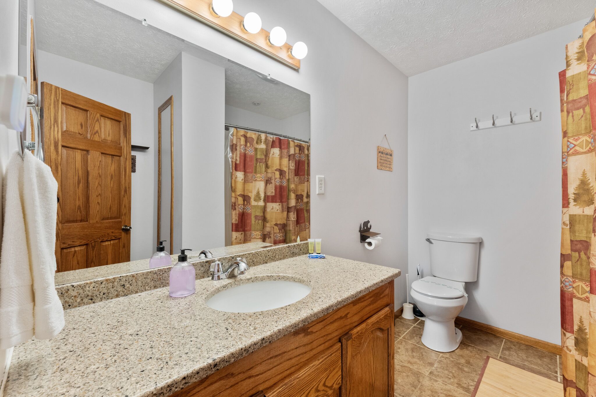 A bathroom with a granite countertop sink, a mirror with lights, a wooden door, a toilet, and a shower curtain.