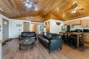 A spacious living area with a wooden ceiling, leather sofas, a round coffee table, and a kitchen with wooden cabinets and a refrigerator.