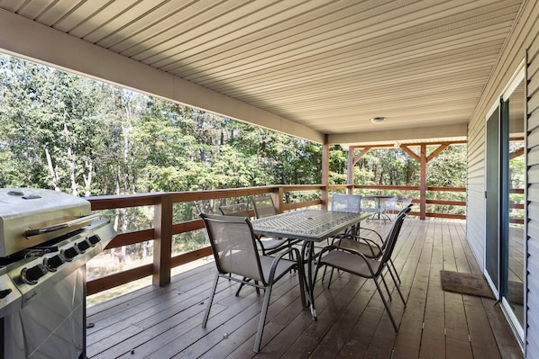 A covered outdoor deck with a grill, metal table, and chairs, surrounded by a wooden railing and overlooking a forested area.