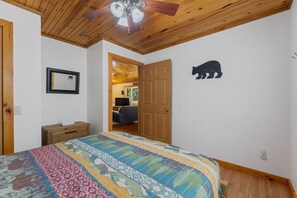 Une chambre avec un plafond en bois, un lit recouvert d’une courtepointe colorée, une table de chevet en bois, un tableau encadré et un autocollant représentant un ours sur le mur.