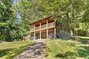 A two-story wooden cabin with a balcony, surrounded by trees and a paved driveway. - Beartastic Outlook | Hocking Hills, Hot Tub (Rockbridge)