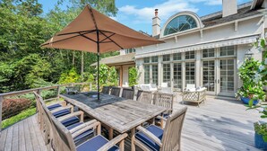 A wooden deck with a table and chairs, a large umbrella, and a view of a house with large windows and a chimney. - Spacious & chic Southampton estate with heated pool, fire pit &... (Southampton)