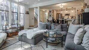 A spacious living room with a grey sofa, a glass coffee table, and a white fur throw. A kitchen is visible in the background with a dining area. - Bright, beautiful condo in the heart of Mammoth, near shuttle, shops, ski & golf (Mammoth Lakes)